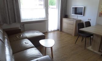 A cozy living room with a brown sofa, a small table, and a television on the wall. Large windows provide bright light in the room.