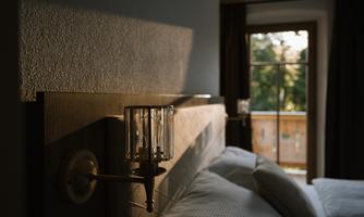A cozy bedroom with a modern headboard and elegant wall lights. The window lets in soft light and a view of the outdoors.