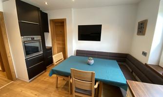 A modern kitchen corner with a table, chairs, and a kitchen unit. The walls are light and the floor is made of wood.