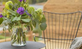A beautiful bouquet of purple and green flower blooms in a glass vase is sitting on a small table. In the background, a comfortable chair and a wooden wall can be seen.