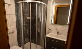A modern bathroom with a shower, a sink, and a mirror. The walls are tiled and there is bright lighting.