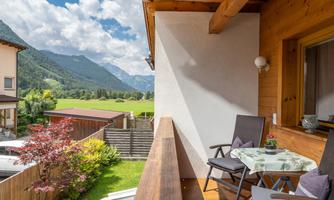 A beautiful balcony with two armchairs and a table. In the background, green mountains and a blue sky can be seen.