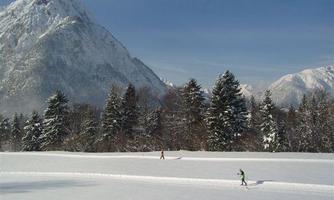 A wintry landscape with snow-covered mountains in the background. Two people are walking on a cross-country ski trail through the sparkling snow cover.