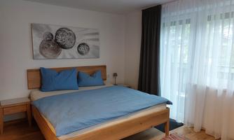 A modern bedroom with a wooden bed and blue pillows. Large windows with curtains provide plenty of daylight.