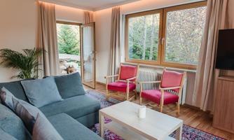 A modern living room with a comfortable sofa and two red chairs. Large windows provide a view of the exterior and plenty of daylight.