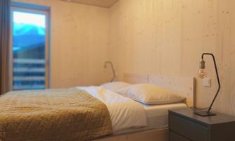 A modern bedroom with wooden walls and a large window. The bed is cozy, with white pillows and a gold-colored blanket.
