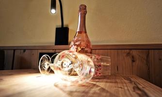 A bottle of rosé sparkling wine stands on a wooden table next to an overturned wine glass. The image is lit by a warm light.