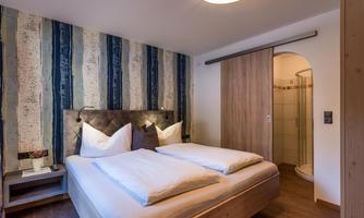 A cozy bedroom with a double bed and two pillows. The walls are decorated with a striped design, and a bathroom with a shower is visible.
