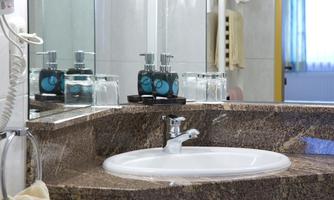 A modern bathroom with a white sink and an elegant granite surface. Toiletries are placed on the shelf, and light reflects through the window.