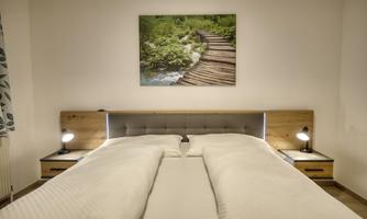 A modern bedroom with a double bed and a light duvet. On the wall hangs a picture of a natural path through a green landscape.