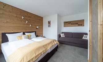 A modern bedroom with a large bed and a cozy sofa. The walls are partially made of wood, creating a warm atmosphere.