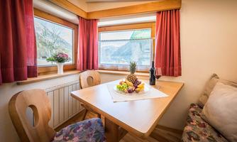 A cozy dining area with a wooden table and chairs. On the table sits a fruit basket and a bottle of wine, while the windows provide plenty of light.