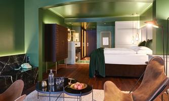 A modern hotel room with green walls and elegant furnishings. The room features a large bed, a comfortable armchair, and stylish decoration.