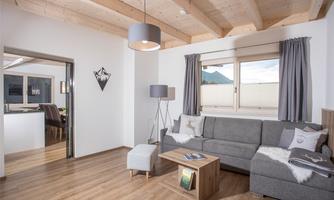 A modern living room with a gray sofa and a table. Natural wooden floor and large windows create a bright atmosphere.