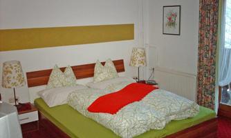 A cozy bedroom with a double bed and colorful pillows. The walls are painted light, and the room has a balcony with a view outside.