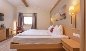 A modern hotel room with two beds, a television, and a cozy armchair. The room features large windows with curtains and a warm, inviting design.