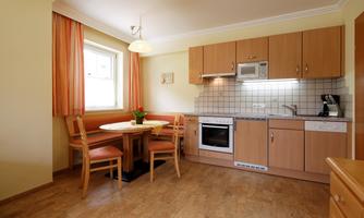 A bright kitchen with wooden cabinets and a round table. The decor is friendly and welcoming, with windows and curtains in warm colors.