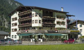 A charming hotel with balcony plants and an inviting restaurant area. The surroundings are characterized by green meadows and mountains.