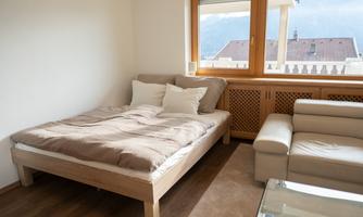 A cozy bedroom with a wooden bed and beige bed covers. Next to the bed is a bright sofa and large windows provide daylight.