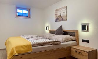 A cozy bedroom with a double bed and a wooden nightstand. A picture hangs on the wall, and a window lets in natural light.