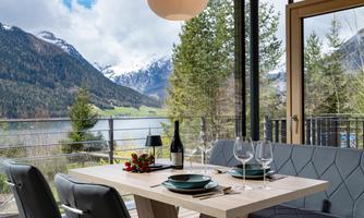 A modern dining room with a view of the mountains and a lake. The table is set with plates, glasses, and a bottle of wine.