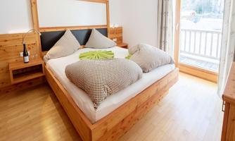A cozy bedroom with a wooden bed and large pillows. The room has a bright ambiance and access to a balcony.