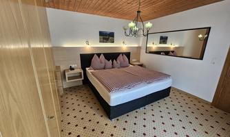 A cozy bedroom with a large bed and wooden ceiling. The room is designed to be bright and friendly, with carpet flooring and decorative elements.