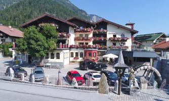 A charming house with balconies adorned with colorful flowers. Several cars are parked in the parking lot, and the surrounding landscape is dominated by mountains.