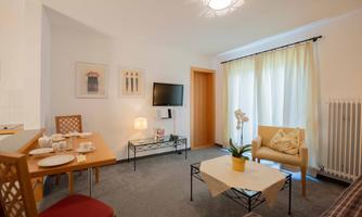 A cozy living room with a dining table, a television, and a comfortable armchair. Large windows let in plenty of daylight, and the decoration is modern and inviting.