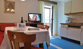 A cozy dining area with a table and chairs. In the background, there is an open kitchen and a television.