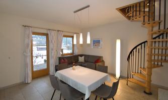 A modern living room with a dining table and comfortable chairs. A spiral staircase leads upstairs, and large windows offer a view of the surroundings.