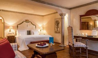 An elegant hotel room with two beds, a wooden table, and cozy furnishings. The room is well-lit and features inviting details.