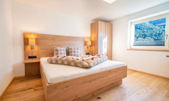 A modern bedroom with a large bed and wooden details. Large windows provide a view of nature.