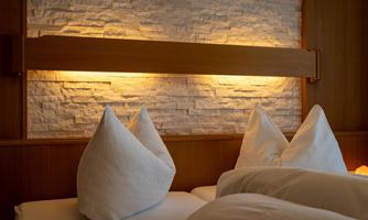 A cozy bedroom with a wooden bed and pillows in classic shape. The illuminated stone wall creates a warm atmosphere.