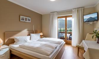 A bright bedroom with a double bed and large windows. The room has a balcony overlooking the mountains and is equipped with a small table and a television.