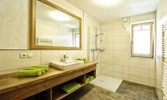 A modern bathroom with a large mirror and a bright shower cabin. On the wooden shelf, there are green towels and the floor is tiled.