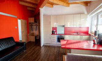 A modern kitchen with red accents and wooden ceilings. In the room, there is a black sofa and light furniture.