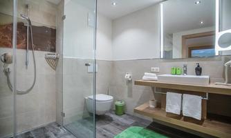 A modern bathroom with a glass shower and a simple sink. The walls are brightly decorated, and there are green accents with towels and care products.