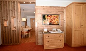 A cozy room with wooden furniture and a television on the wall. In the background, a dining area with chairs and a table is visible.