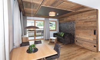 A modern living room with wooden decor, a dining table, and a cozy couch. Large windows provide a view of the terrace and plenty of daylight.