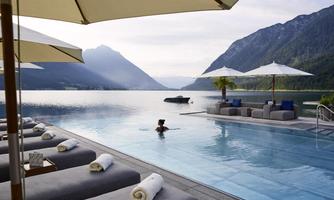 A luxurious pool area with comfortable loungers and parasols, surrounded by mountains and a clear lake. A person swims relaxed in the pool.