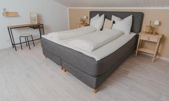 A modern bedroom with a large gray bed and white pillows. Additionally, there is a small desk and a bright, friendly atmosphere.