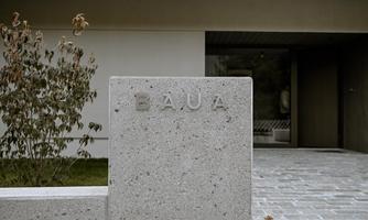 A modern entrance area with a grey stone column on which the word "BAU" is engraved. In the background, plants and a clear entrance area can be seen.