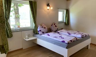 A bright bedroom with a large bed and colorful pillows. Large windows let in a lot of light and offer a view outside.