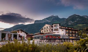 A picturesque hotel in the mountains with a scenic landscape in the background. The buildings are surrounded by green plants and a dramatic sky.