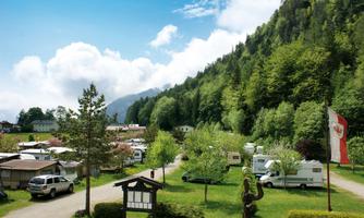 A campsite surrounded by green trees and hills. Motorhomes and cars are parked on the grounds, and the sky is clear and blue.