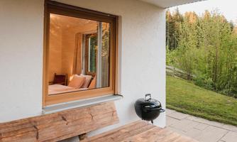 A cozy terrace with a wooden bench and a grill. Through the window, one can see a bright room with a bed and green nature in the background.