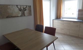 A simple dining area with a wooden table and chairs. The walls are light, and there is a view of the garden through the window.