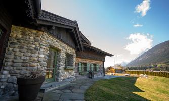 A traditional stone house with large windows overlooking a green meadow. In the background, mountains and a clear sky can be seen.