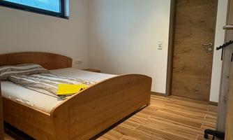 A simple bedroom with wooden flooring and a large bed. The room has a window and a wooden door.
